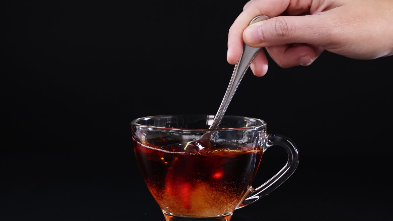 A hand stirs sugar into a glass cup of tea against a black background, creating a calming visual effect