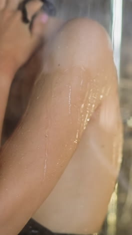 young woman puts back under hot flowing water streams in fogged shower cabin reflecting daylight at morning daily routine slow motion closeup