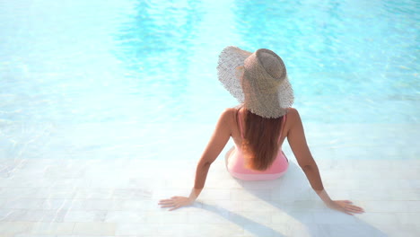 Back-view-of-motionless-woman-with-large-hat-and-pink-swimsuit-sitting-in-pool-water