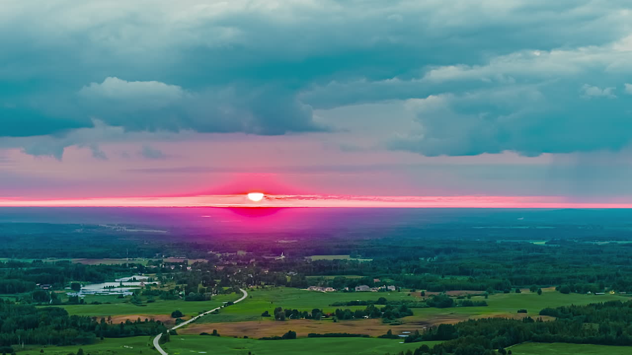 A stunning aerial timelapse captures a vibrant pink and purple sunset over the vast rural landscape of the Latvian countryside, showing forests, fields, and a small village as sun rolls over horizon