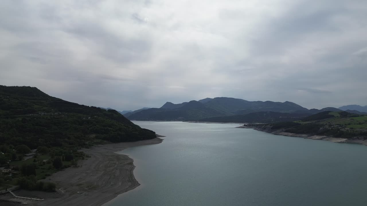 Smooth 4K drone panning footage capturing the side of Lac de Serre Ponçon dam with cloudy skies, surrounding trees, water, and mountains. Scenic aerial view in the French Alps, May 2025