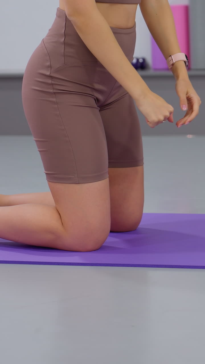 vista parcial de una mujer en traje deportivo marrón arrodillada en una alfombra púrpura en un gimnasio moderno, otra persona en rosa es parcialmente visible en primer plano, alfombras de ejercicio y equipos de fitness