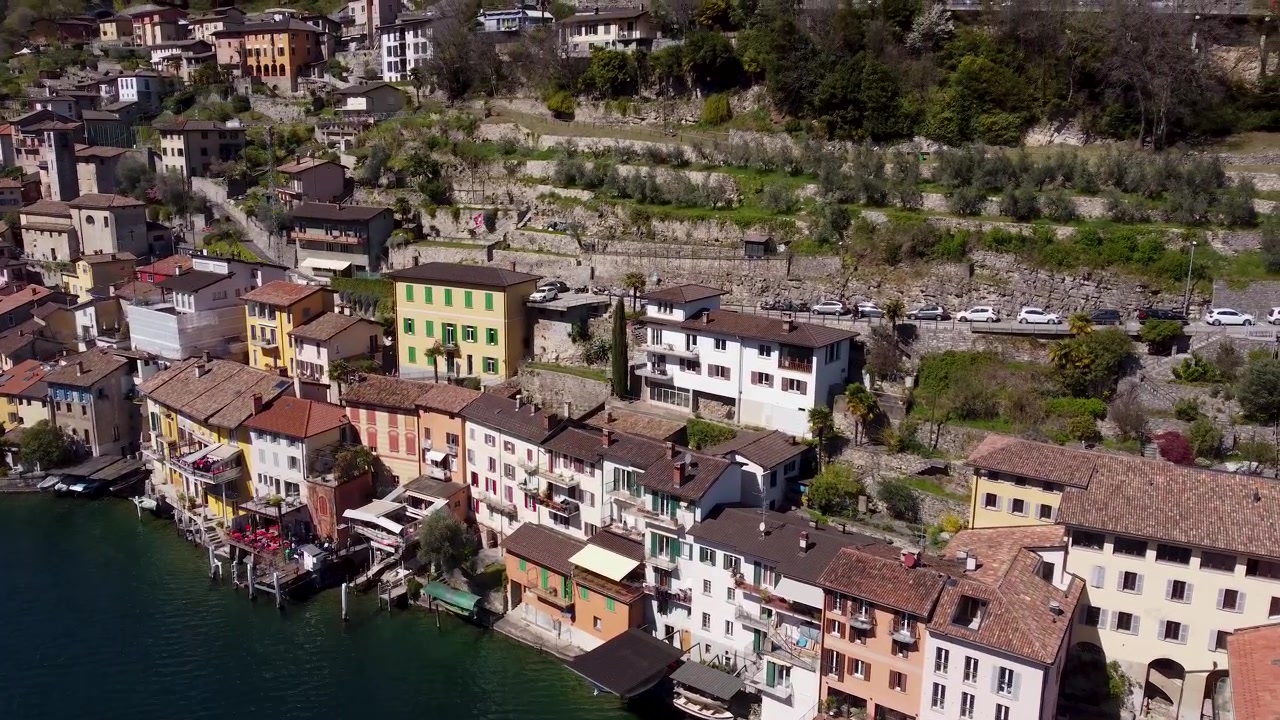 Case colorate in riva al lago si aggrappano a un ripido pendio terrazzato a Gandria, vicino al Lago di Lugano, Svizzera; piccole barche di legno attraccano lungo il lungomare vicino a terrazze di ulivi verdi. Ripresa aerea orbitale con drone.