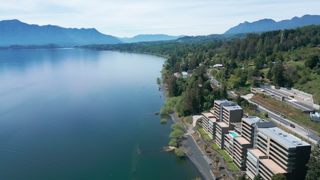 Backward Drone Shot of Serene Lake and Winding Road Surrounded by Lush Forest and Distant Mountains on a Clear Day