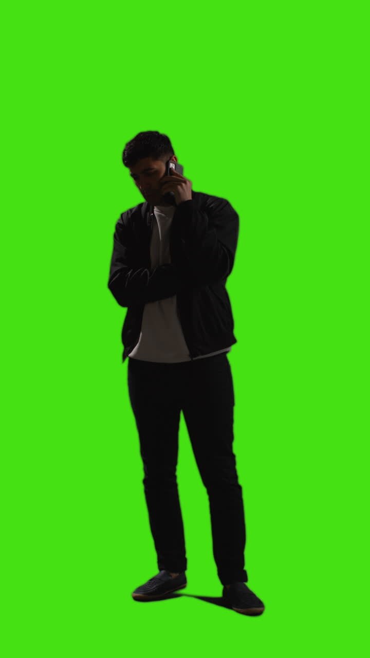 Video verticale, inquadratura a figura intera di giovane uomo che parla al telefono cellulare in piedi davanti a uno schermo verde con illuminazione a bassa chiave 2