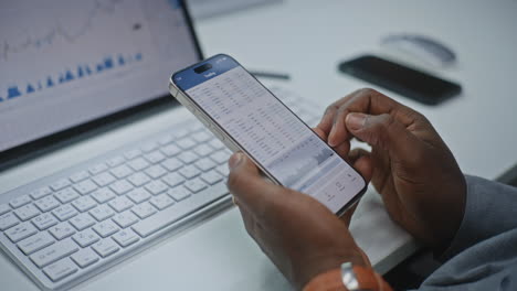 african american financial analyst checking real-time stocks, exchange market charts on smartphone and digital tablet using online trading app. businessman working in investment bank office. close up.