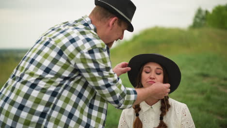 an artist in a checked shirt adjusts the hat of a woman with braided hair, wearing a white dress, seated in a green field, smiling shyly