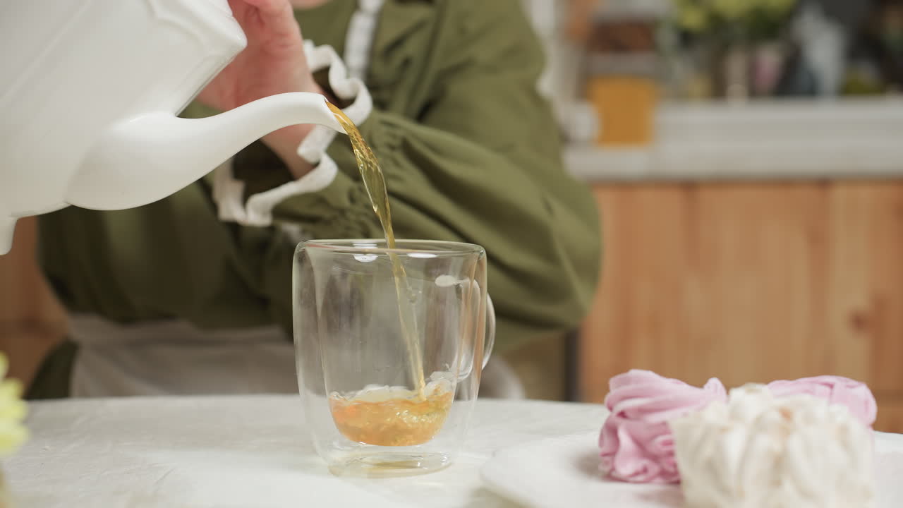 Close up of woman in green dress pouring hot coffee from white ceramic teapot into transparent glass cup on white tablecloth with sweet meringue dessert in background