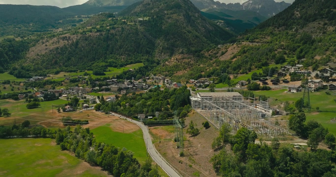 Un drone si allontana da una sottostazione elettrica ad alta tensione con importanti linee di trasmissione, rivelando una spettacolare valle alpina circondata da maestose montagne