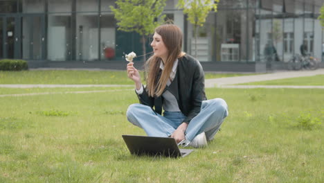 young stylishly dressed girl sitting in the park on the grass near the computer holding a dandelion