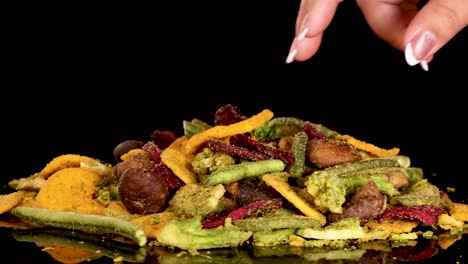 a hand reaches into a colorful pile of assorted dried vegetable chips, picking up a piece under bright studio lighting against a black backdrop. camera remains steady