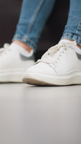 primer plano de las zapatillas blancas de lona de la dama tocando suavemente el pie izquierdo y derecho en el suelo con pantalones de jean mientras está sentado en un sofá en un entorno relajado de un centro comercial