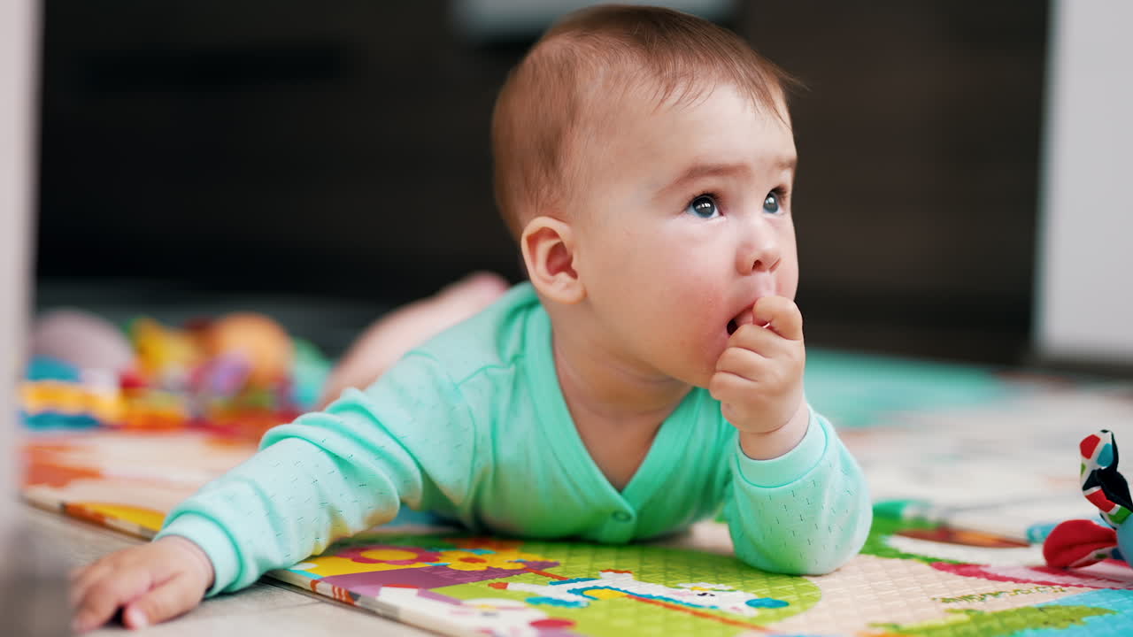 Lovely little kid lying on the colorful mat and looking up. Sweet toddler putting his thumb into mouth. Blurred backdrop.