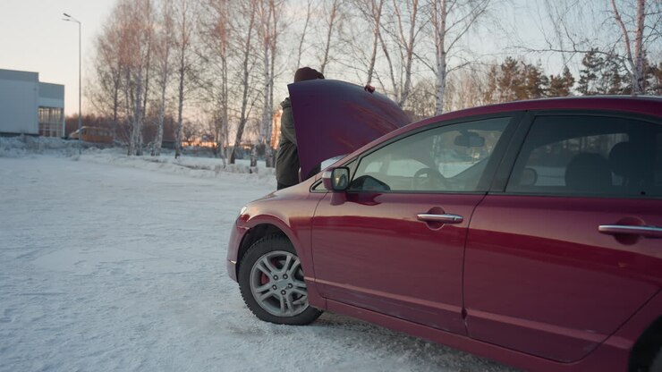 Man in winter jacket opens bonnet of parked red car and inserts rod support to hold it open for inspection or maintenance, with snowy road, leafless trees, and distant building