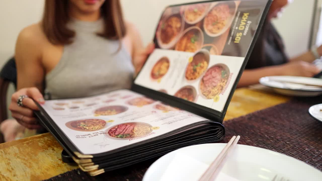 A woman in a sleeveless top flips through a food menu at a restaurant table, with another diner in the background. Natural indoor lighting, shallow depth of field