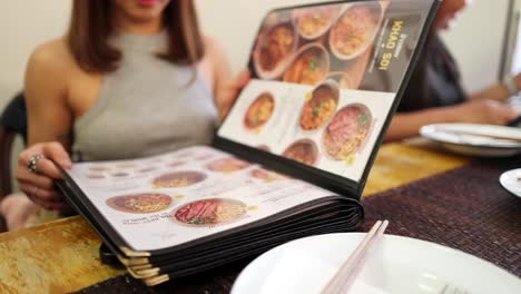 a woman in a sleeveless top flips through a food menu at a restaurant table, with another diner in the background. natural indoor lighting, shallow depth of field