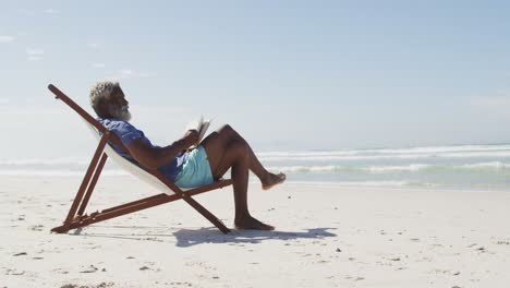 hombre afroamericano mayor leyendo y acostado en una cama de sol en una playa soleada