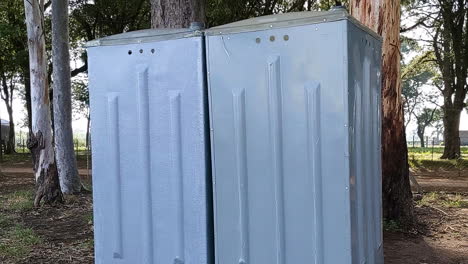 Handheld-close-up-shot-of-grey-portable-bathroom-surrounded-by-trees-in-public-nature-setting