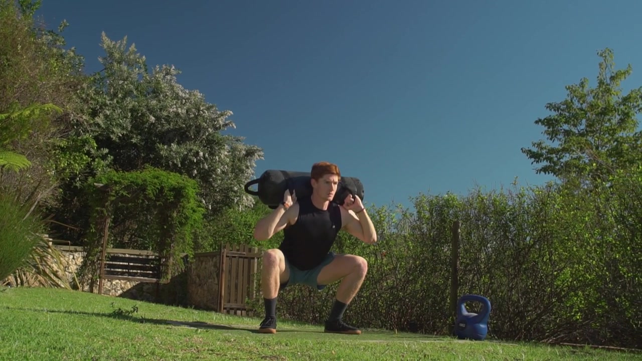 Giovane uomo atletico con i capelli rossi tiene un pesante sacco di sabbia nero sulle spalle mentre esegue squat profondi su un prato. Inquadratura ampia statica dal basso.