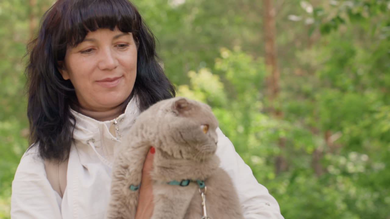 Smiling Female Embraces Relaxed Gray Feline Outdoors, Cheerful Woman Lovingly Holds Her Calm Gray Cat In Park, Joyous Woman Comfortably Cuddles Her Serene Gray Feline Amid Lush Green Foliage Outdoors