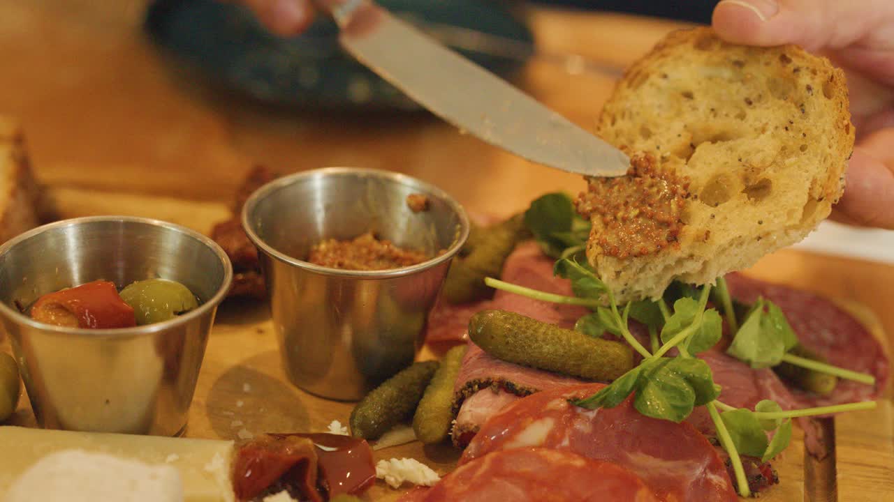 A hand spreads pâté onto a slice of rustic bread above a charcuterie board with cured meats, cheese, pickles, and microgreens in warm indoor lighting