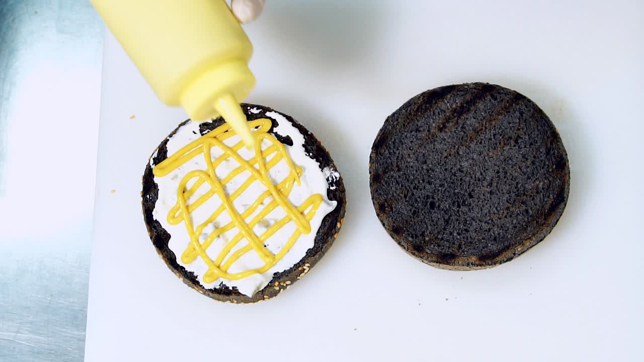 Making black burger. A cook spreading mustard on a burger bun on white background. Preparation of vegan black hamburger. Cooking tutorial. Close-up
