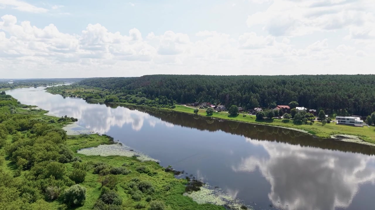 Spettacolare video con drone che sorvola un fiume tranquillo costeggiato da foreste verdeggianti e case di campagna, sotto un cielo azzurro brillante con nuvole soffici che si riflettono sulla superficie dell'acqua.