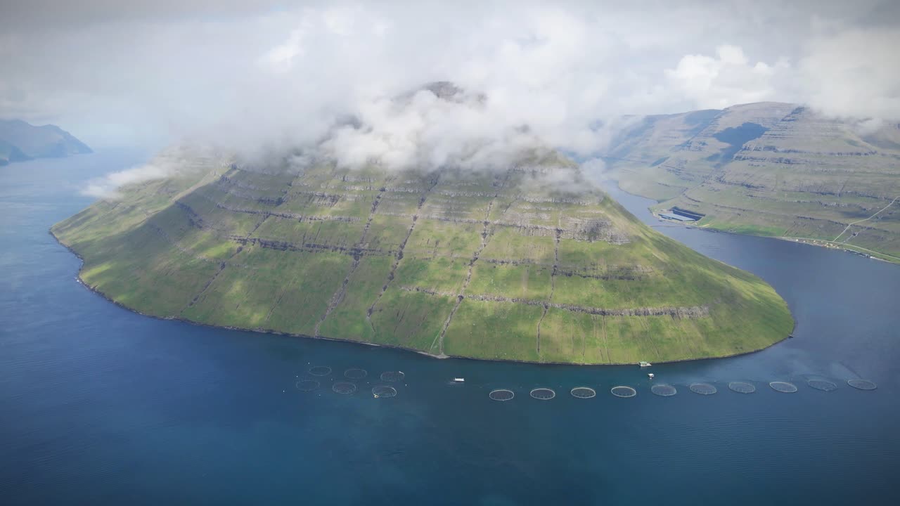 페로 제도 (faroe islands) 의 클라크스비크 (klaksvik) 근처에 있는 연어 농장을 공중에서 찍은 사진으로, 거친 산과 조용한 물과 조화롭게 번성하는 수산업이 강조됩니다.