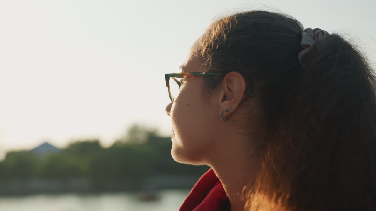 Young woman gazing lakeside at sunset, wearing glasses and red hoodie, ponytail catching golden backlight, calm water with distant boats, soft cinematic glow, contemplative mood and relaxed posture