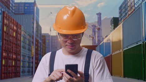primer plano de un trabajador asiático con gafas de protección y casco de seguridad sonriendo y usando un teléfono inteligente mientras está de pie en un almacén de contenedores