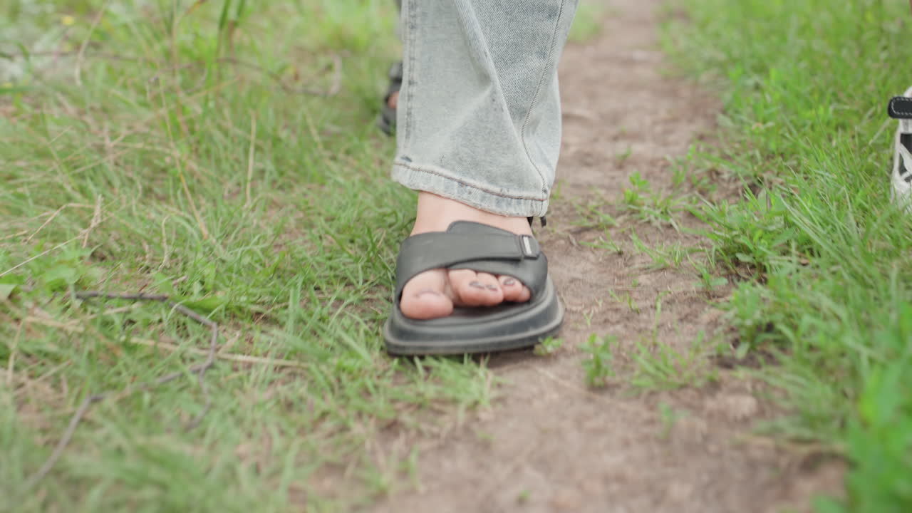 Walking sandals through natural terrain, Adult in jeans carefully stepping on grassy dirt pathway footage, Detailed view of sandals on dirt trail with calm earthy ambiance and leisurely pace
