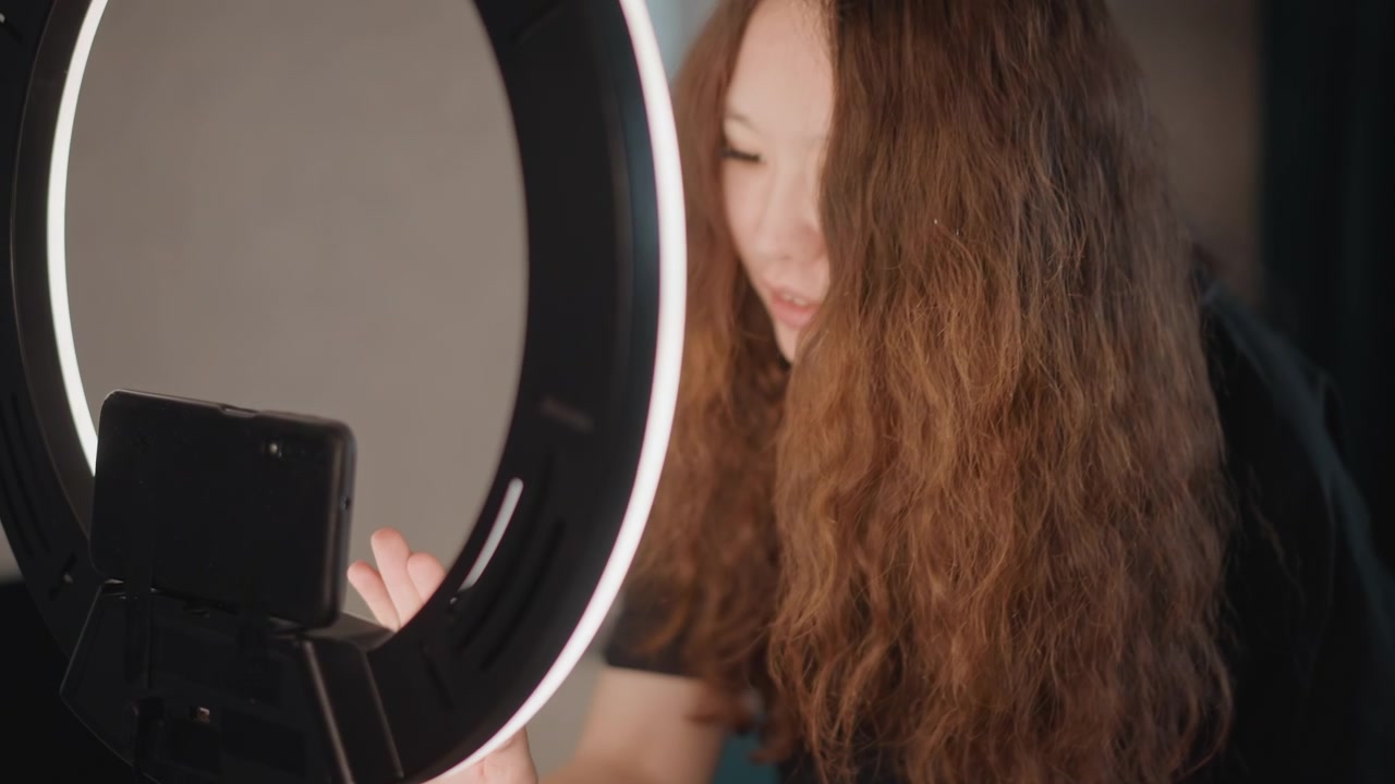 Mujer joven se inclina hacia adelante hacia una luz anular LED iluminada y ajusta el soporte del smartphone con una inclinación de cabeza y movimiento de mano visibles, su cabello largo y ondulado ocultando parcialmente el rostro.