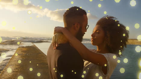 animation of spots of light over happy diverse couple embracing by sea