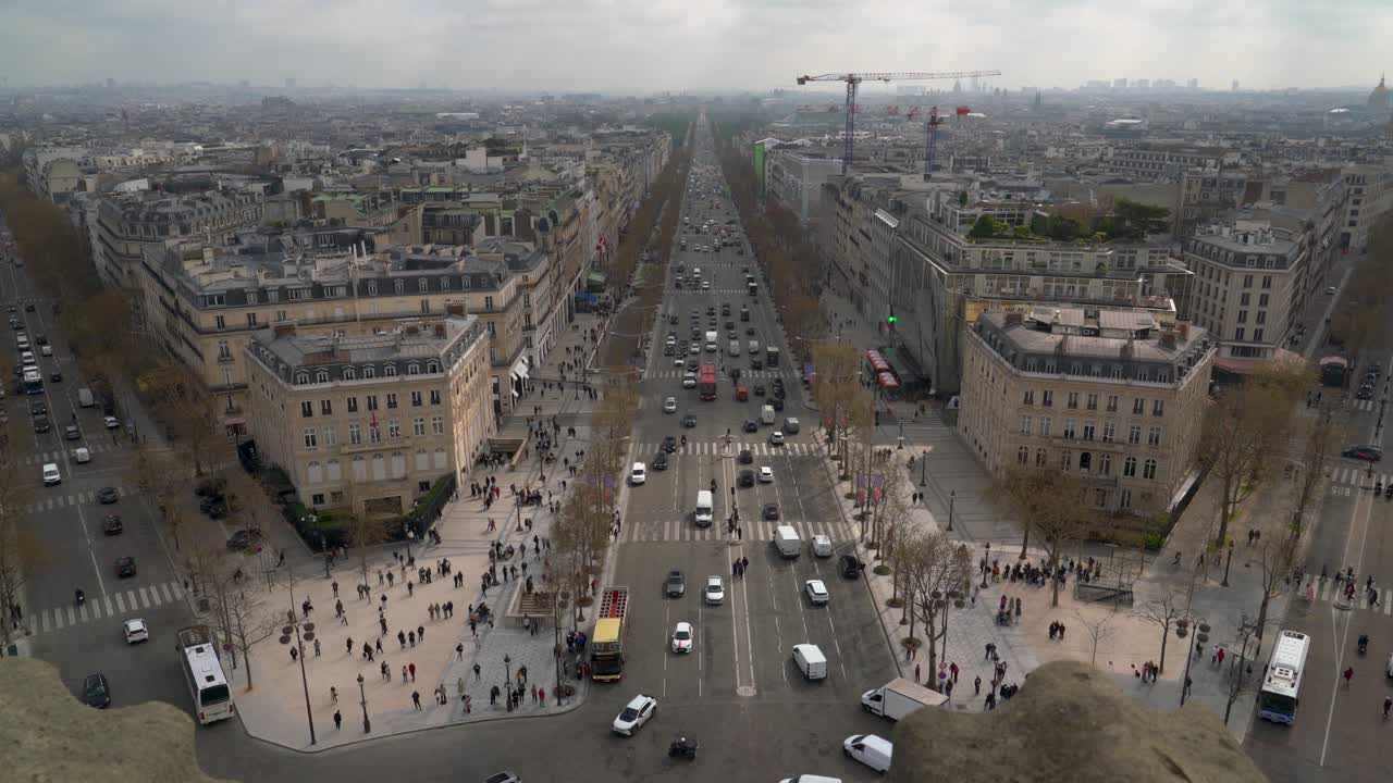 Looking out over Paris from the top of the Arc de Triomphe intersection in the heart of the city of love while traffic and tourists go by along the city streets below on a cloudy day