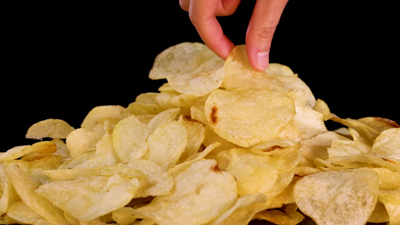 Human hand reaches into pile of potato chips, brightly lit, isolated on black background, close-up