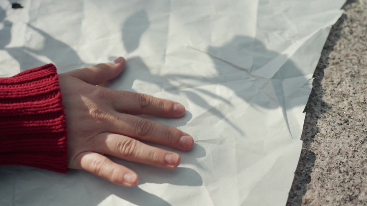 Primo piano di una mano con manica rossa che tocca e leviga carta bianca stropicciata su una superficie di pietra texturizzata, proiettando un'ombra dal fogliame vicino. La carta mostra pieghe e texture mentre la luce solare crea interessanti giochi di luce.