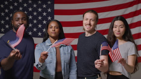 Studio-Portrait-Shot-Of-Multi-Cultural-Group-Of-Friends-Waving-Miniature-Stars-And-Stripes-Flags-In-Front-Of-American-Flag-Celebrating-4th-July-Independence-Day-Party-1