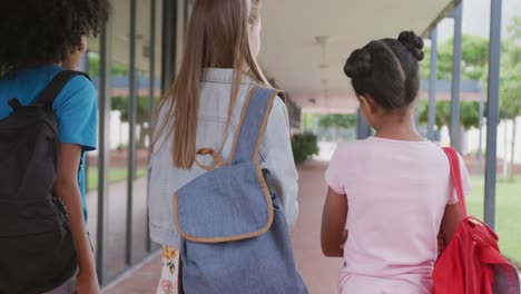 Video-of-rear-view-of-three-diverse-schoolgirls-walking-in-school-corridor-talking,-copy-space