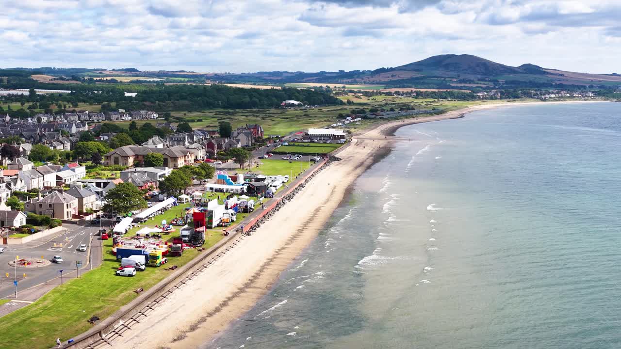 Drone glides above Fife coastline, revealing seaside town, beach, seawall, and distant hills in daylight