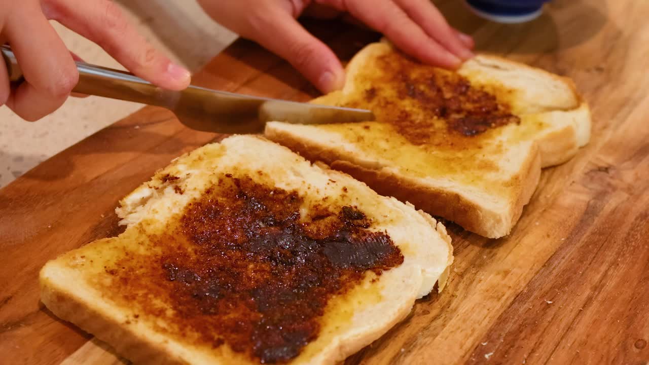 Close-up of hands using a knife to spread a dark spread on toasted bread slices on a wooden board.