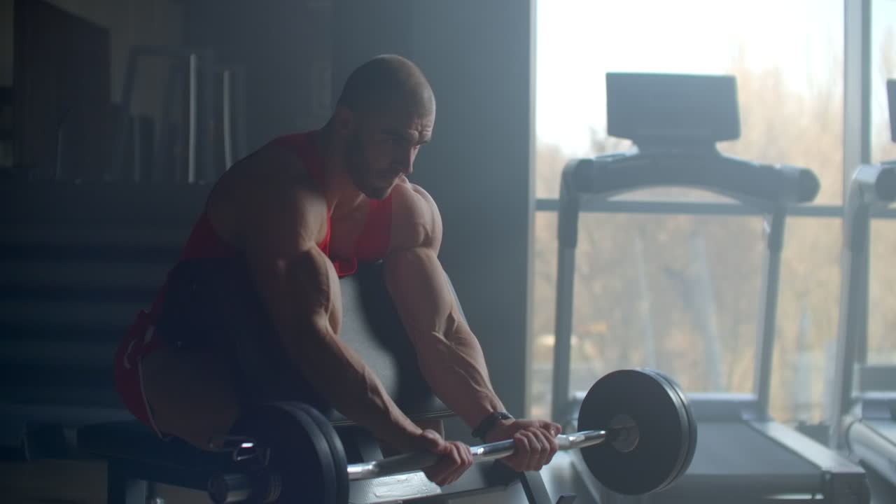 Young strong sweaty man biceps workout training with heavy barbell in the gym dark with shadows real people. Young strong muscular sweaty man with big