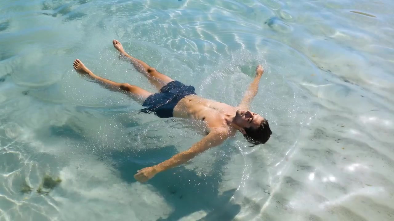 Uomo che galleggia sentendo la deriva, aggiustando gli arti mentre rimane in equilibrio in acqua bassa, con costume da bagno scuro.