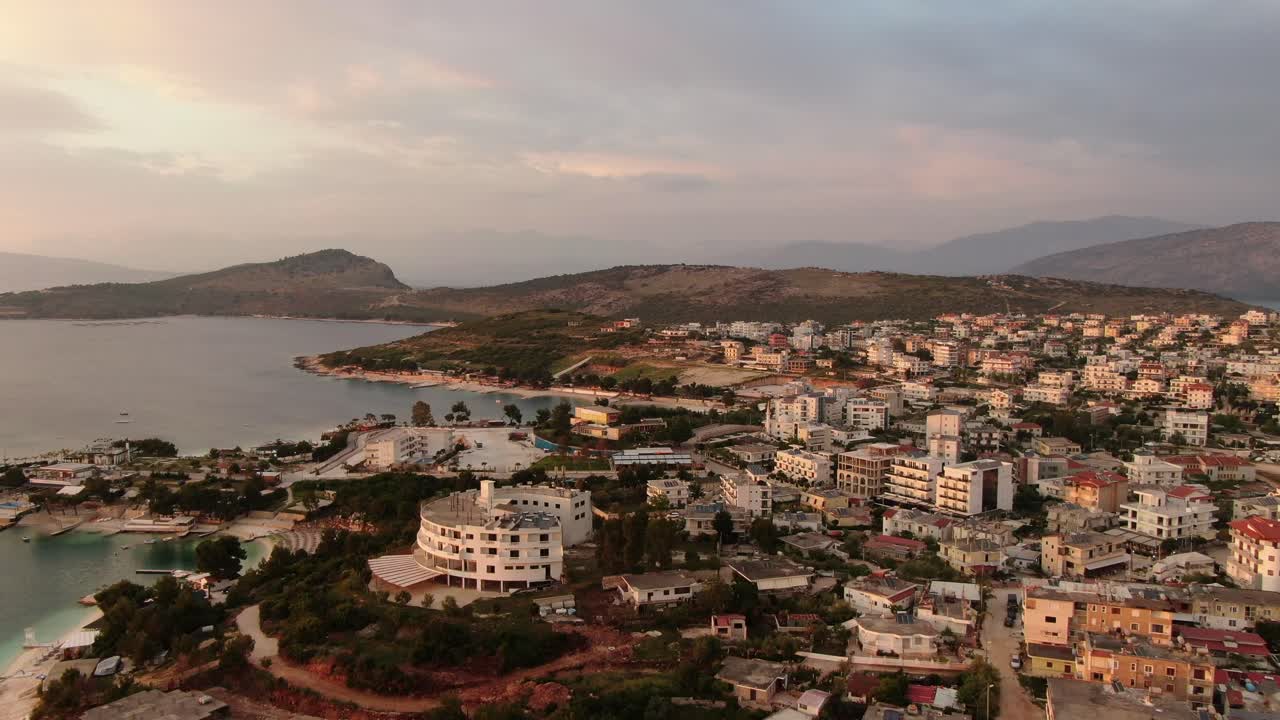 vista de avión no tripulado en albania volando sobre aguas cristalinas azules al atardecer, hoteles y edificios blancos con colinas verdes en ksamil