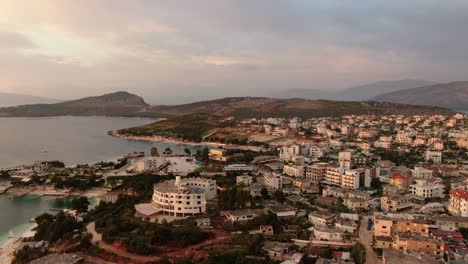 Drone-view-in-Albania-flying-over-blue-crystal-clear-water-on-sunset,-hotels-and-white-buildings-with-green-hills-in-Ksamil