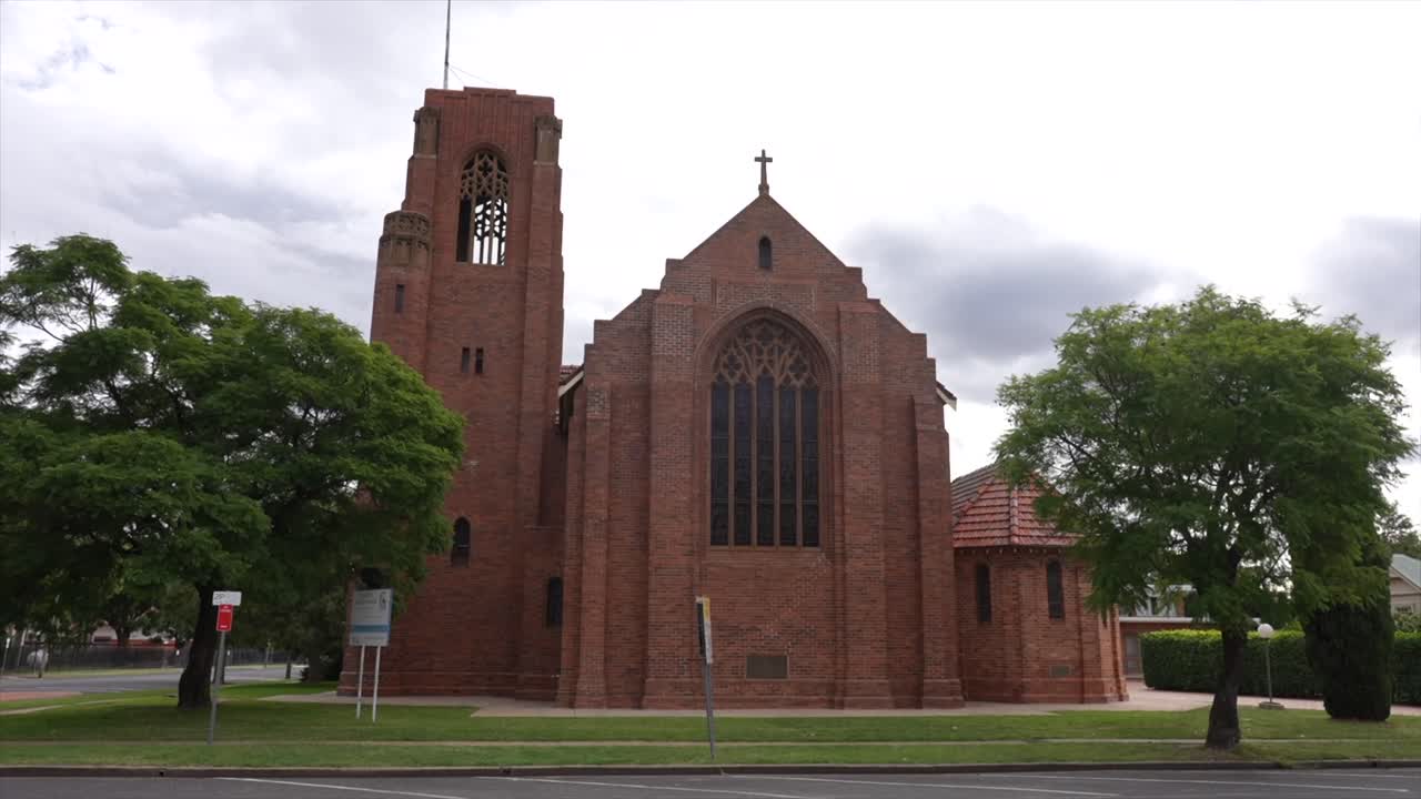 Wide static shot of a historic red-brick church in Moree, New South Wales. The scene shows the tall bell tower, arched windows, cross on the roof, and surrounding green trees under an overcast sky