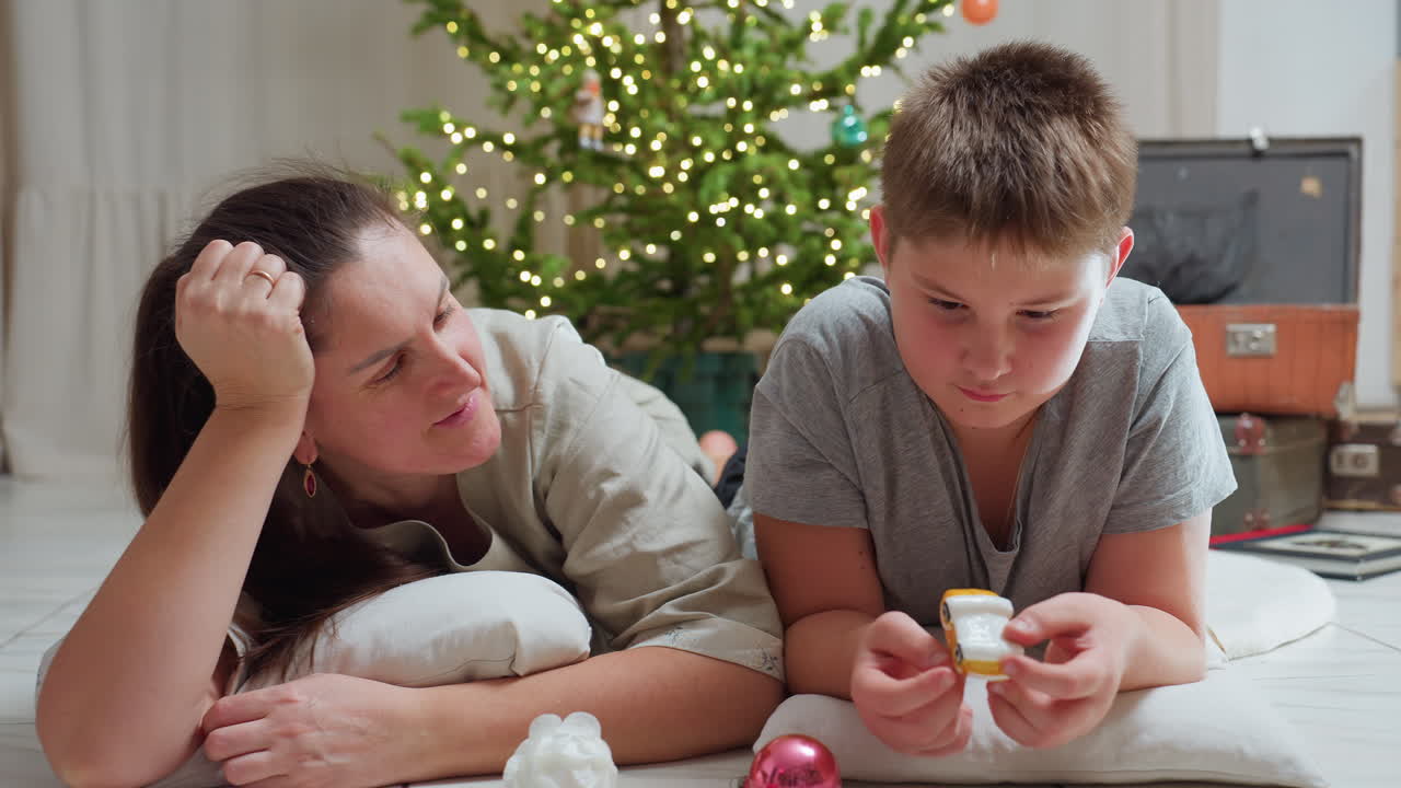 mother and son lying on marble floor using pillows boy holding colorful christmas ornament cozy indoor setting background featuring decorated christmas tree festive atmosphere suitcase nearby