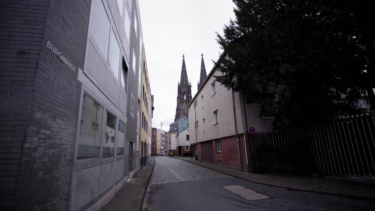 A quiet street in Cologne leads the eye to the iconic twin spires of the Cologne Cathedral, rising above the urban landscape like a timeless monument.