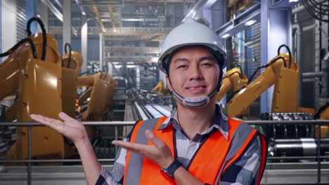 close up of asian male engineer with safety helmet smiling and pointing to side while standing with industrial robotic arms working