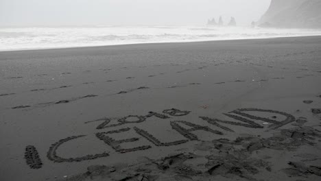 Statische-Aufnahme-Eines-Textes-Aus-Island-2018,-Der-In-Den-Schwarzen-Sand-Geschrieben-Wurde,-Während-Wellen-Schlugen,-Den-Strand-Und-Schwarze-Klippen-Am-Arktischen-Meer,-An-Einem-Stürmischen-Tag,-Am-Diamond-Beach-An-Der-Südküste-Islands