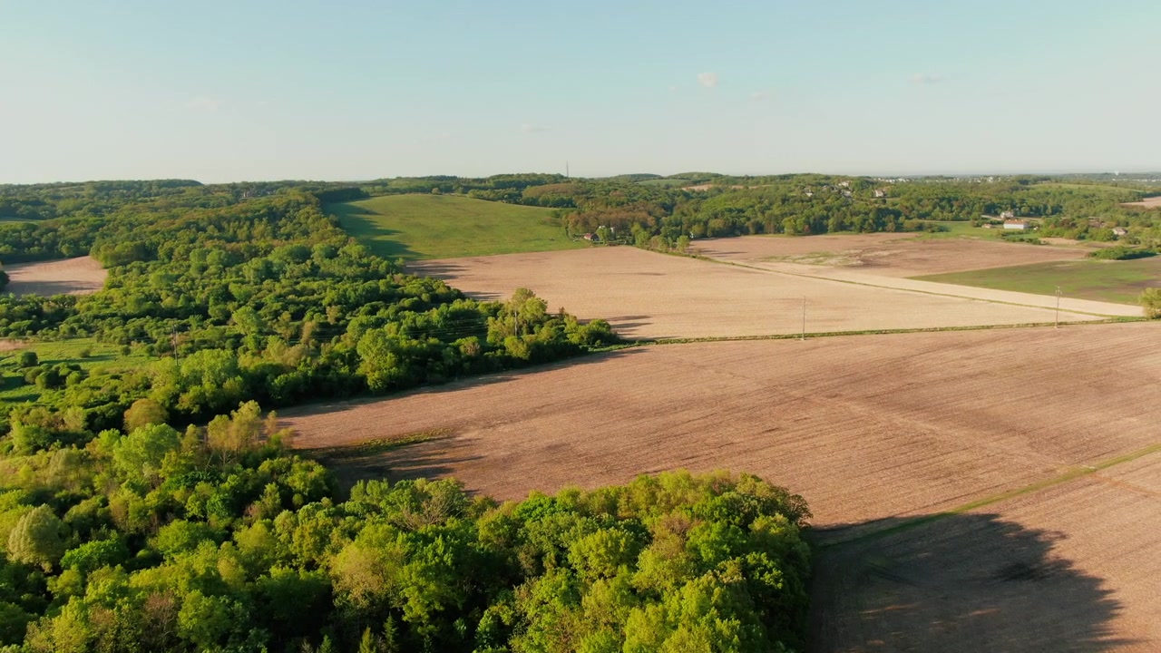 Aerial establishing shot of vast farmland bordered by dense green forests under a clear sky, in Waukesha, Wisconsin. Ideal for agriculture or environmental projects.